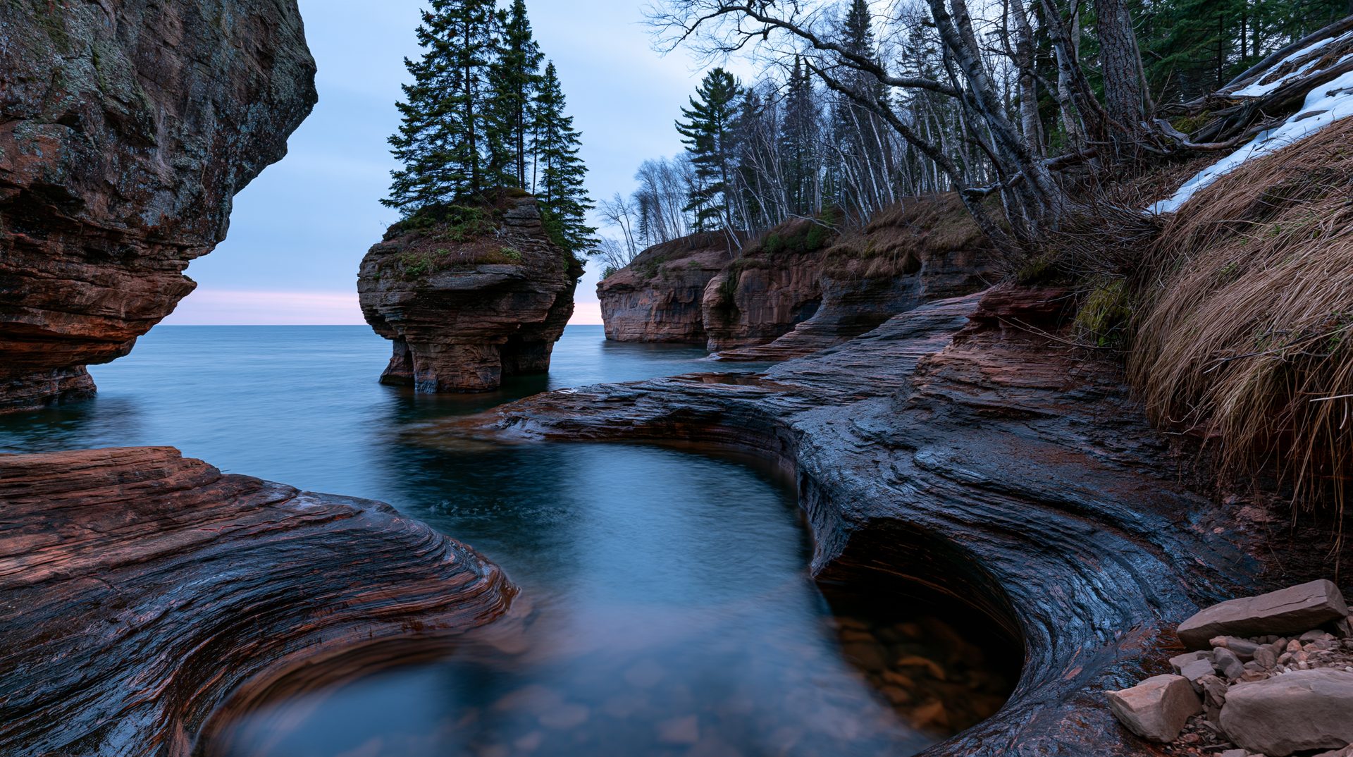 A wide-angle landscape photograph at twilight showing a prominent, tree-topped sandstone rock pillar standing in calm blue lake water, framed by swirling, layered rock channels and cliffs along a rugged coast with remnants of winter snow.