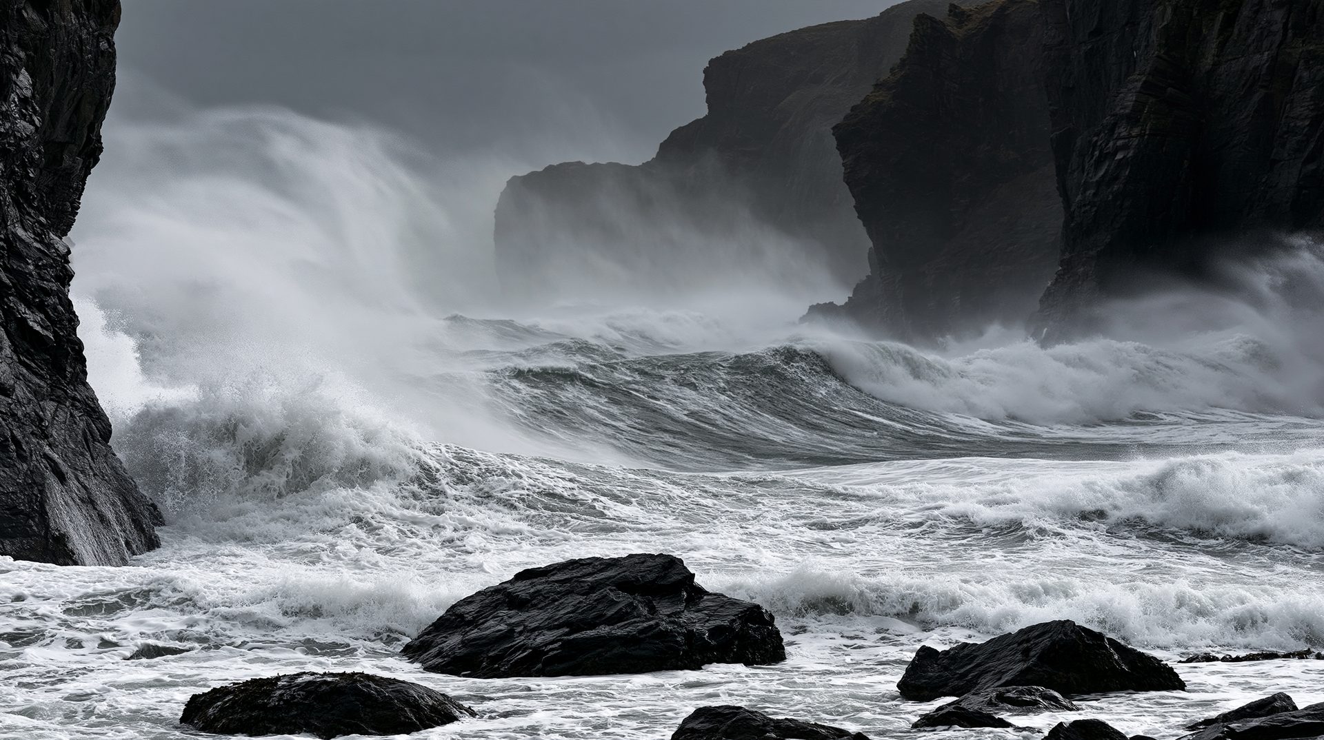 A dramatic black and white photograph of massive, powerful ocean waves crashing violently between dark, steep sea cliffs during a coastal storm.