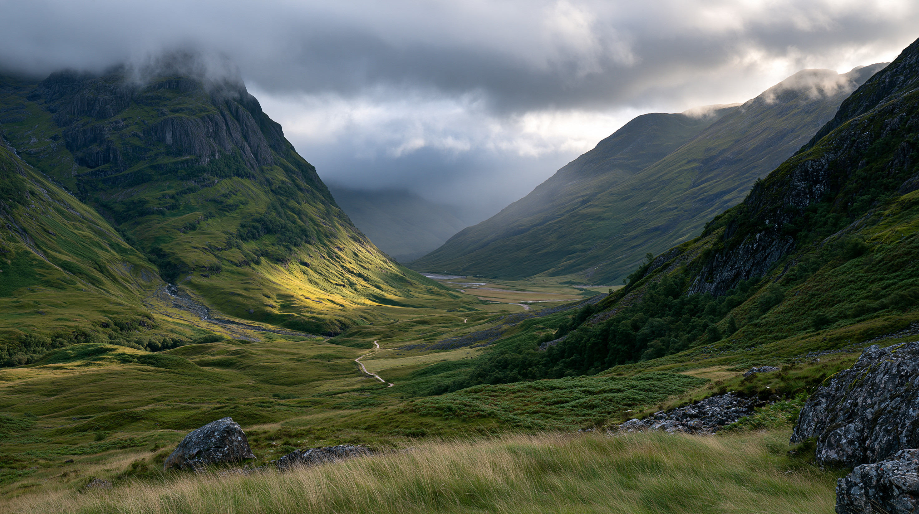 A breathtaking, wide landscape photograph of a deep, green valley in the Scottish Highlands under dramatic storm clouds, with a single patch of bright sunlight illuminating the valley floor.