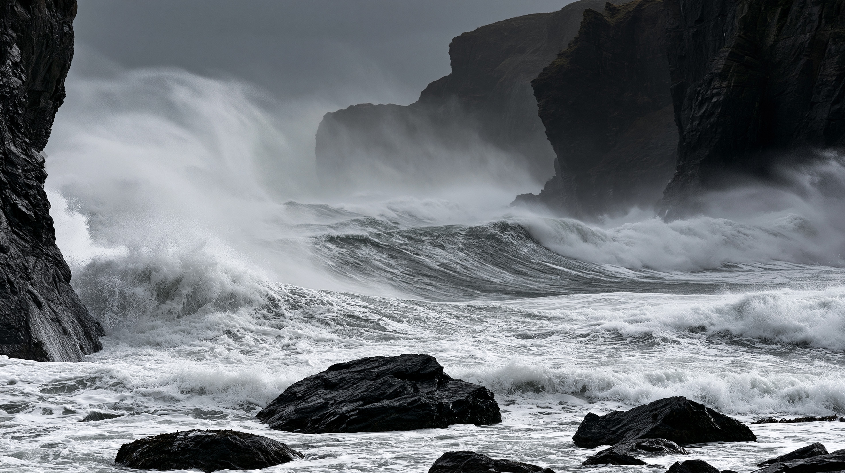 A dramatic black and white photograph of massive, powerful ocean waves crashing violently between dark, steep sea cliffs during a coastal storm.