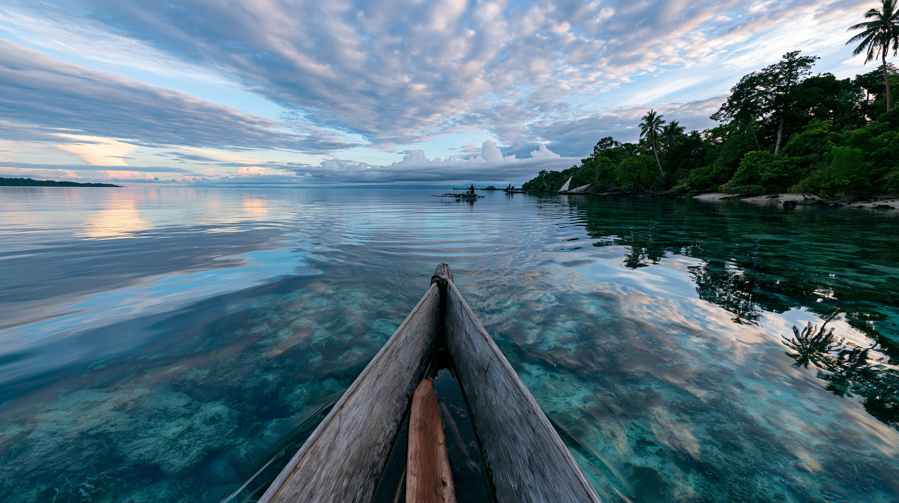 First-person wide-angle perspective from the bow of a wooden dugout canoe gliding over glass-calm, crystal-clear turquoise ocean water revealing a shallow coral reef, with a lush tropical island shoreline on the right under a soft, cloud-filled sunrise sky.