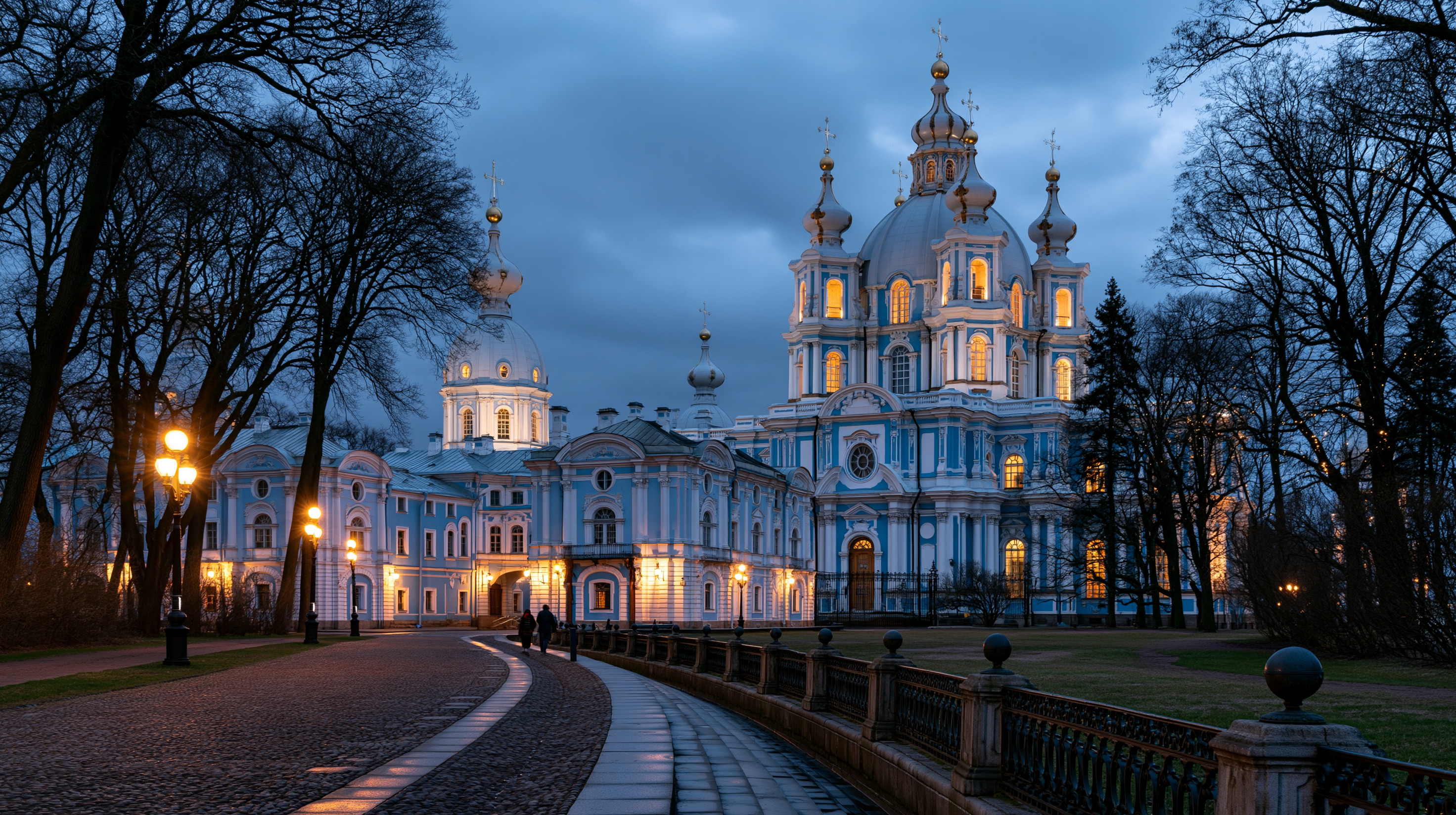 A majestic blue hour view of the illuminated blue and white Smolny Cathedral at the Smolny Convent in Saint Petersburg, featuring a curved cobblestone path, streetlamps, and bare winter trees.
