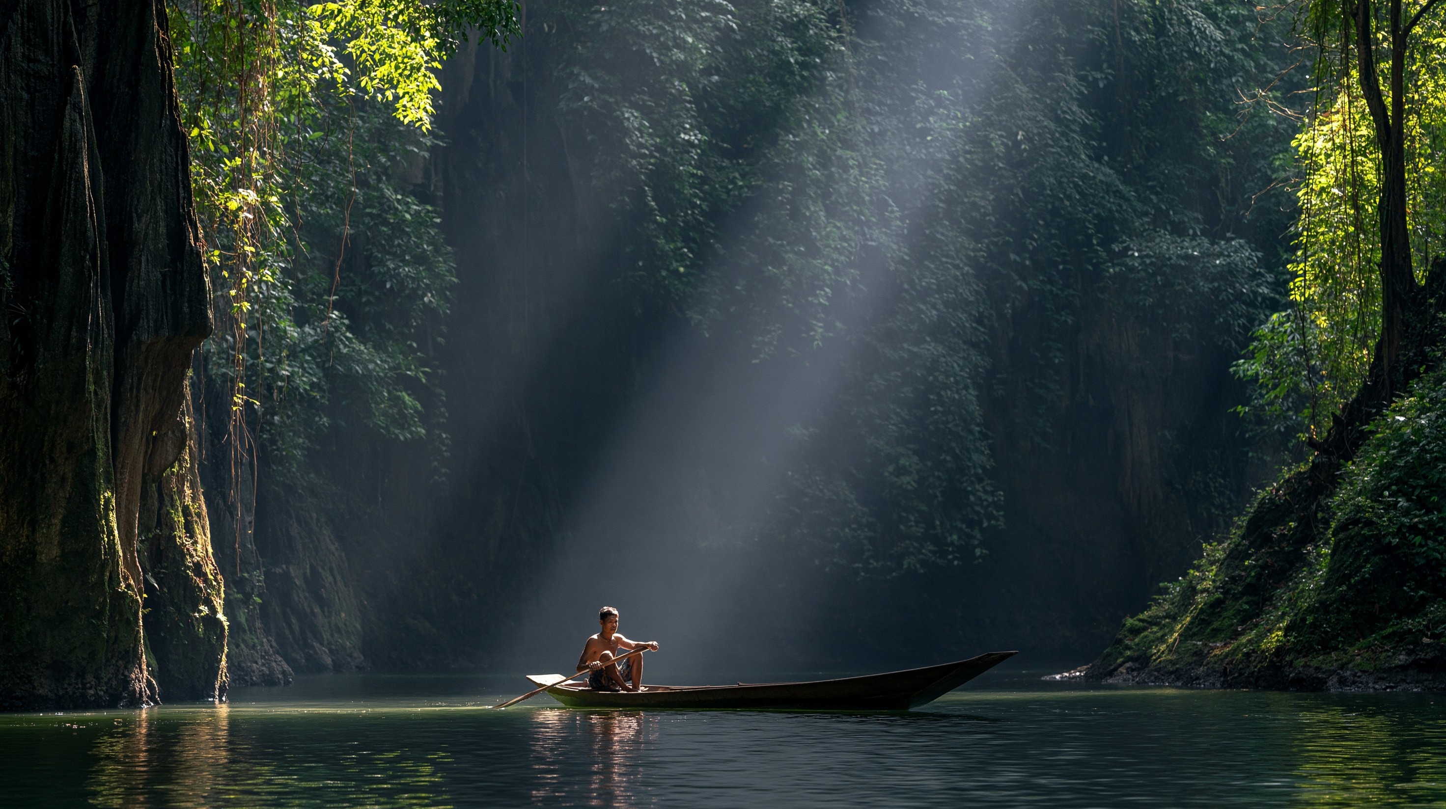 A dramatic landscape view from inside a dark limestone canyon in Khao Sok, Thailand, showing a solitary boatman illuminated by a thick, brilliant sunbeam cutting down through a misty green jungle canopy onto a calm river.