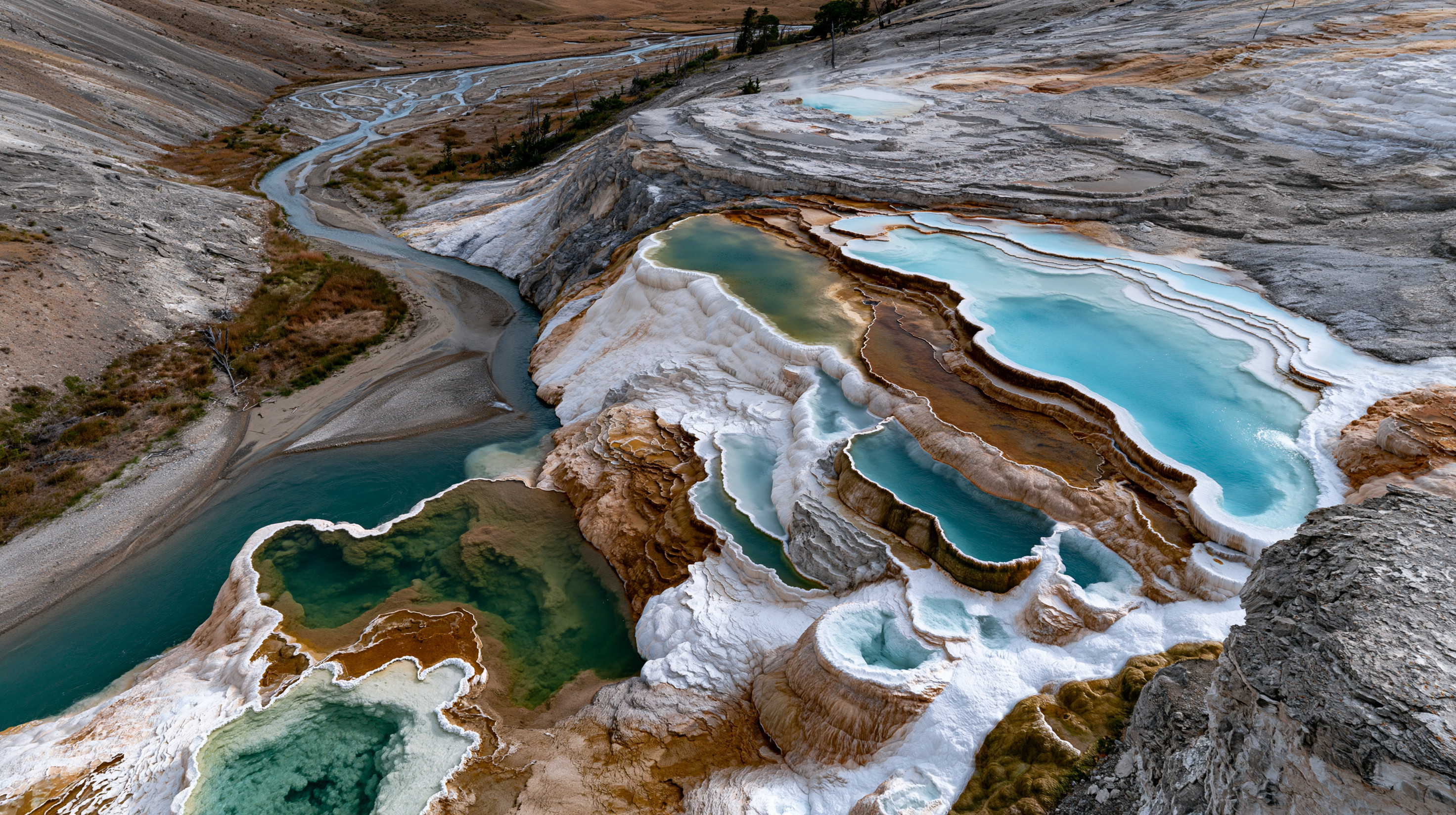 A high-angle landscape shot of the vibrant turquoise and white travertine terraces of Mammoth Hot Springs with a winding mountain river valley visible below.