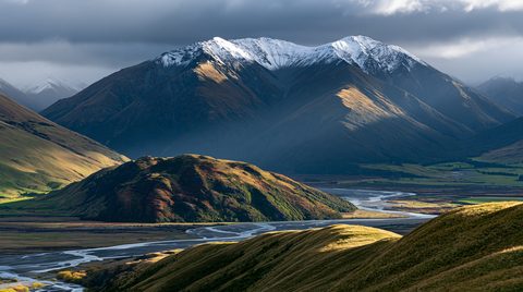 A majestic wide shot of the Ahuriri River valley in New Zealand during a storm, showcasing a braided river winding through a vast valley under dramatic, dark rain clouds with sun rays spotlighting rolling green and yellow hills below snowy mountain peaks.