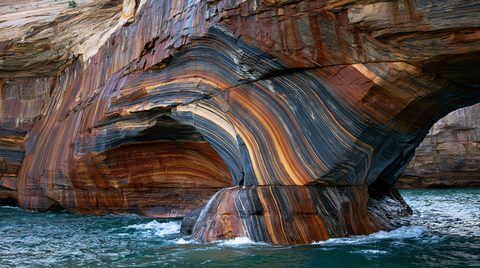 A water-level close-up photograph of a massive, swirling sandstone arch at Pictured Rocks National Lakeshore, showcasing dramatic curved bands of orange iron and black manganese stains with a single cormorant perched on a rock ledge above choppy teal water.