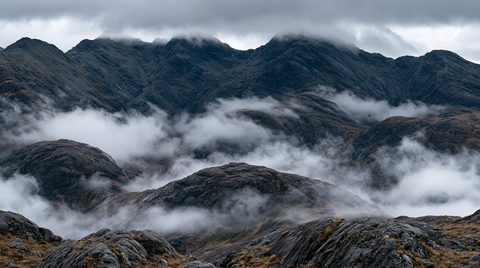 A dramatic, layered landscape photograph showcasing dark, jagged mountains under a heavy overcast sky. Thick swaths of white morning mist drift through the valleys, twisting around rugged, undulating foothills in the foreground and mid-ground. The high, massive rock faces in the background are heavily textured with steep ridges and dark crevasses, their highest peaks partially hidden inside a low-slung ceiling of gray storm clouds.