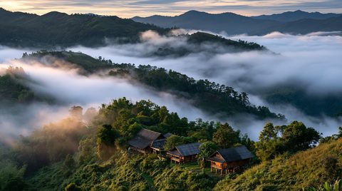 An elevated landscape view of traditional wooden stilt houses on a lush green mountain slope overlooking a deep valley completely filled with thick morning mist under a soft sunrise in Phetchabun, Thailand.