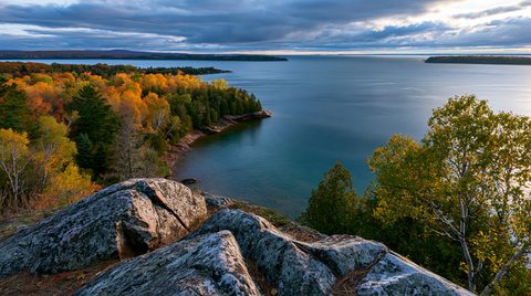 Wide-angle landscape photograph from a rocky granite overlook looking down at a forested peninsula covered in vibrant orange and yellow autumn colors along the blue waters of Lake Superior in Wisconsin.