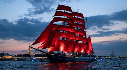 A twilight wide view of the illuminated brig Rossiya with vibrant scarlet sails sailing on the Neva River in Saint Petersburg, Russia, with the golden spire of the Peter and Paul Fortress visible in the background.