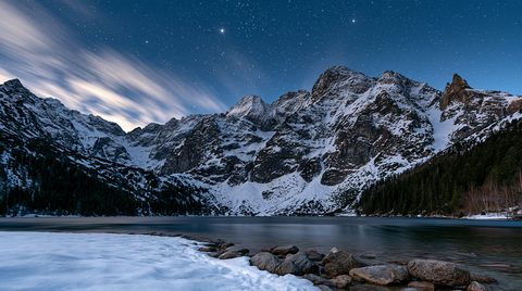 A wide-angle night photograph of a dark mountain lake surrounded by towering, snow-capped granite peaks under a brilliant starry sky with streaks of white clouds.