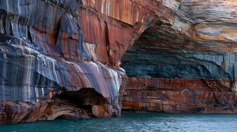 A close-up water-level shot of towering, vibrant sandstone cliffs at Pictured Rocks National Lakeshore, showcasing massive overhanging sea caves stained with deep red iron and black manganese bands, with small cormorants perched on the rock ledges.